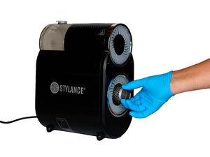 A black STYLANCE countertop appliance with a blue disposable glove covering cleaning a dyson airwrap brush attachment